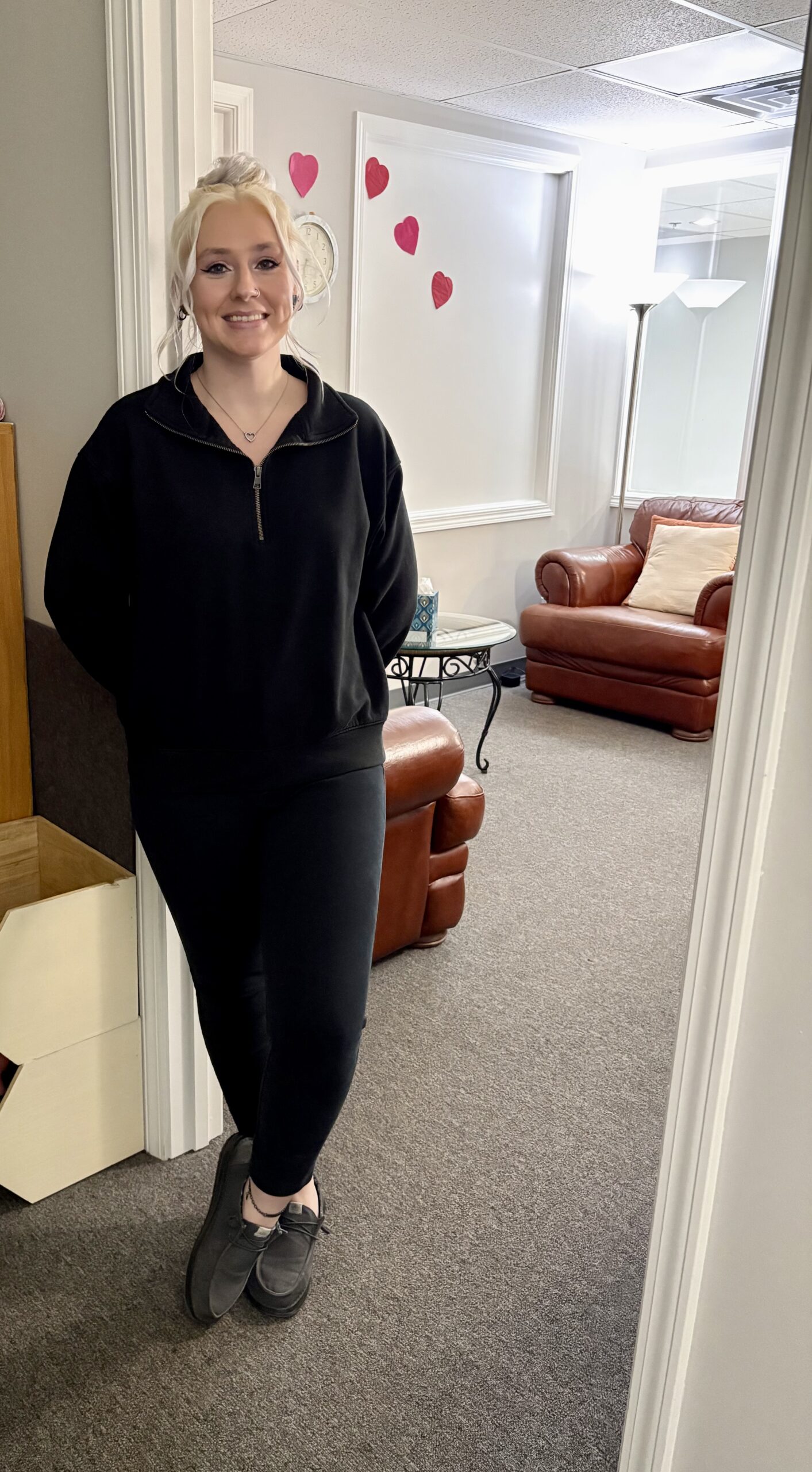 A person with blonde hair, wearing a black outfit, stands and smiles in a doorway. Behind them is a room with brown leather chairs, a lamp, and pink heart decorations on the wall.