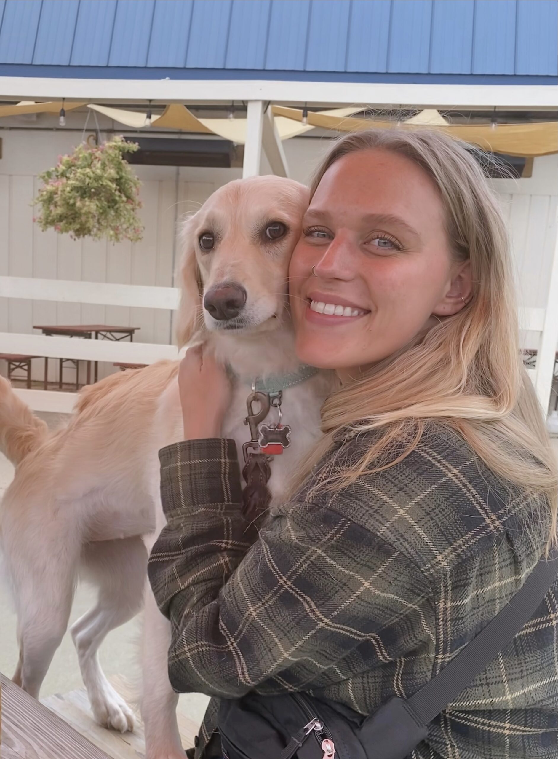 A smiling woman in a plaid jacket hugs a light-colored dog with droopy ears while sitting outdoors at a picnic table. The background features white fencing, wooden benches, and hanging plants.