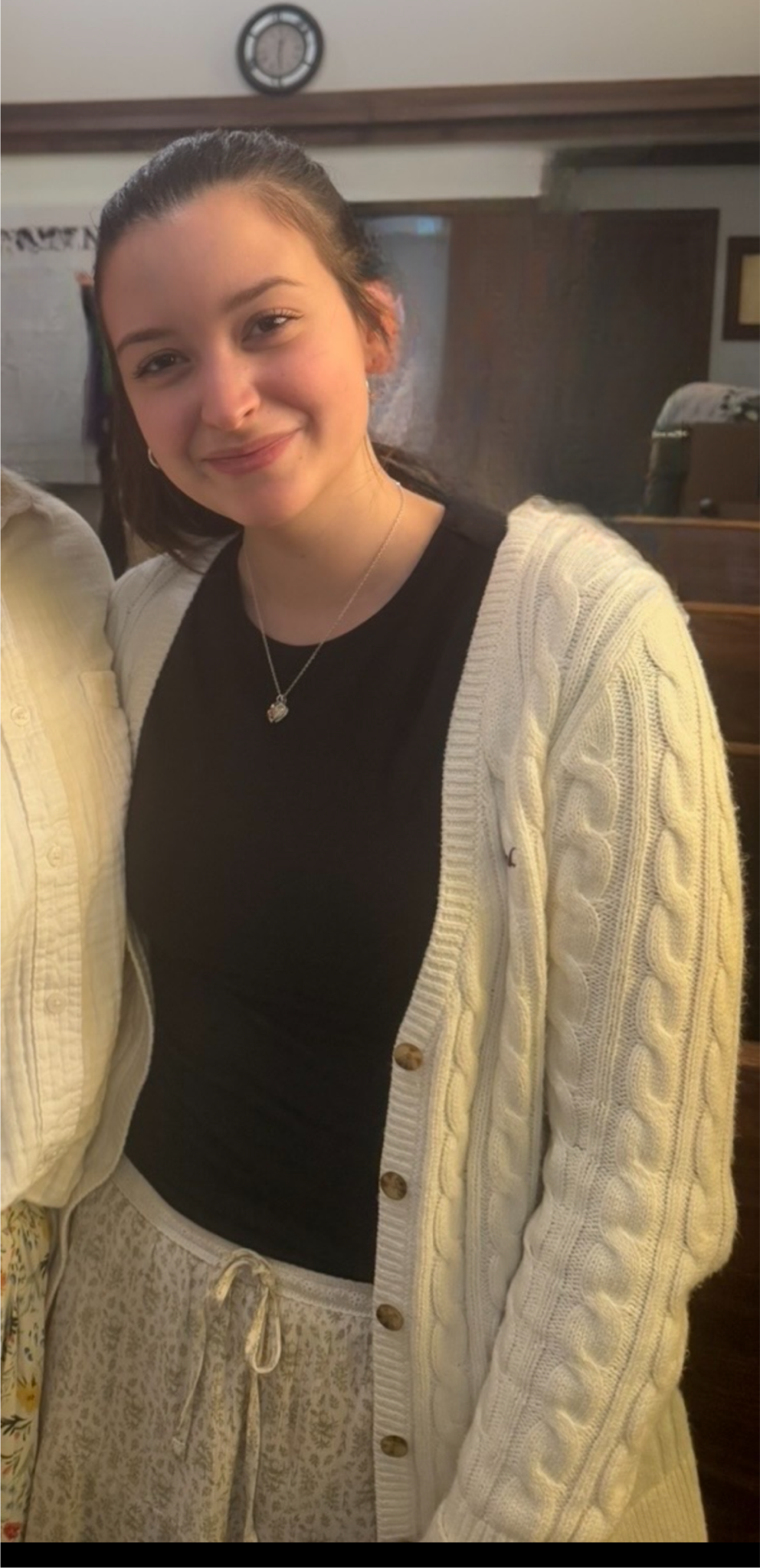 A young woman with long dark hair tied back, wearing a white cable-knit cardigan over a black top and light lace-patterned pants, stands indoors and smiles at the camera.