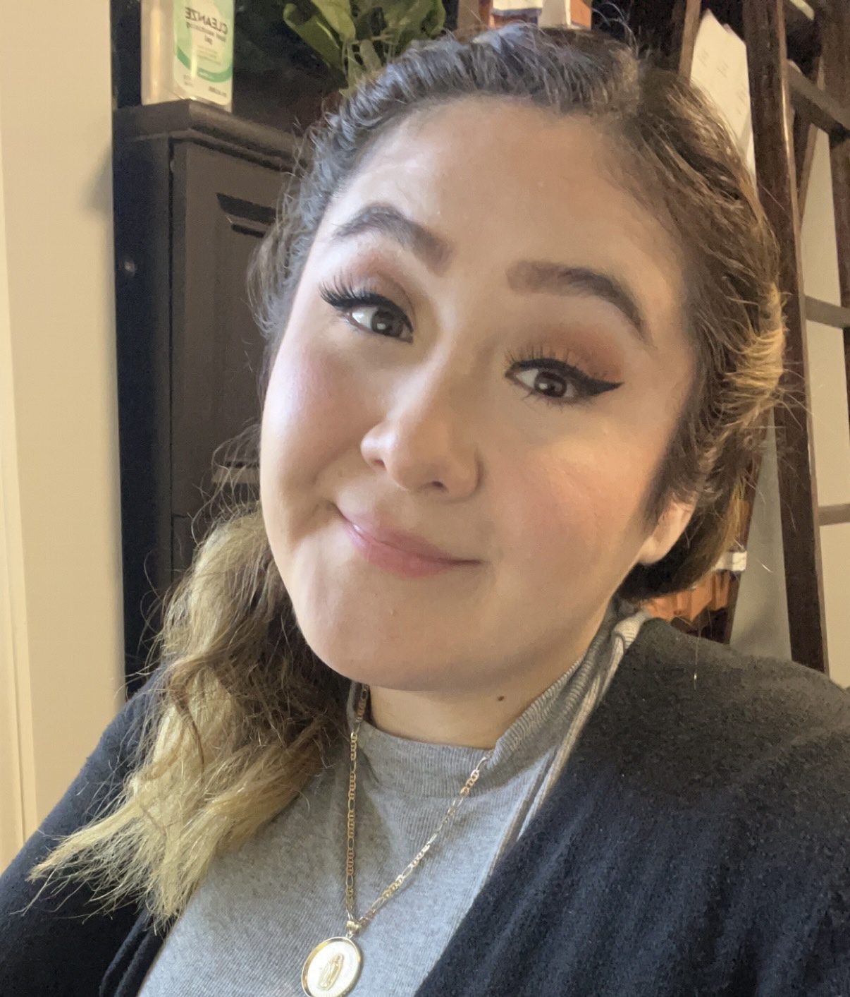 A woman with long brown hair in a side braid is smiling softly at the camera. She is wearing a gray shirt, a dark sweater, and a gold necklace with a pendant. Shelves and a plant are visible in the background.