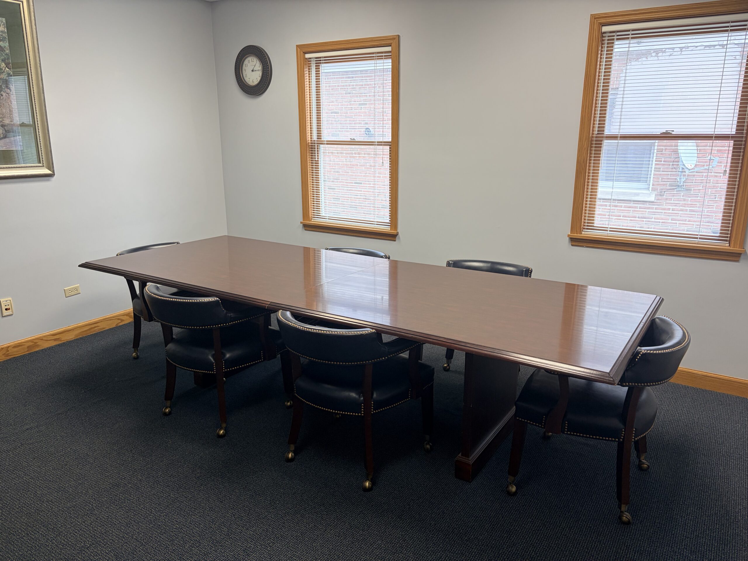 A conference room with a long wooden table surrounded by six black chairs, two windows with blinds, a wall clock, and a framed picture on a light gray wall.