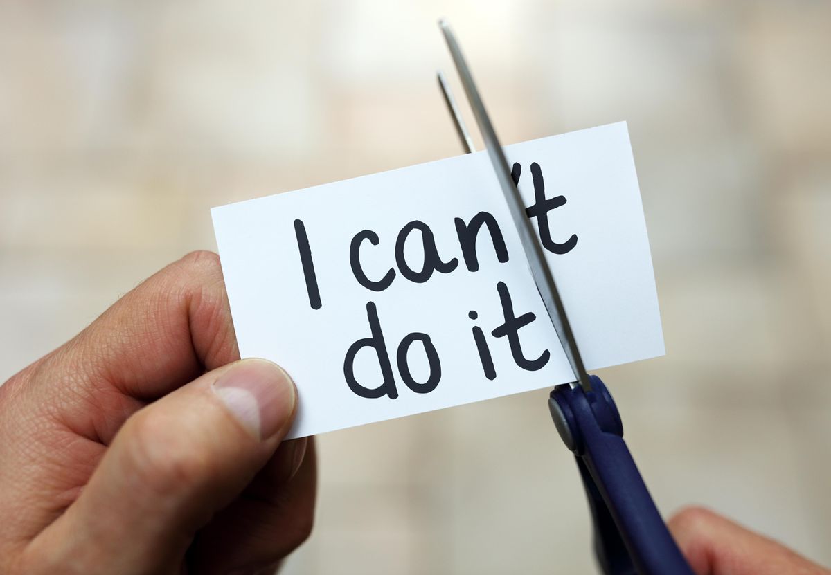 A hand holds a card that says I cant do it while another hand uses scissors to cut off the t, changing the message to I can do it.