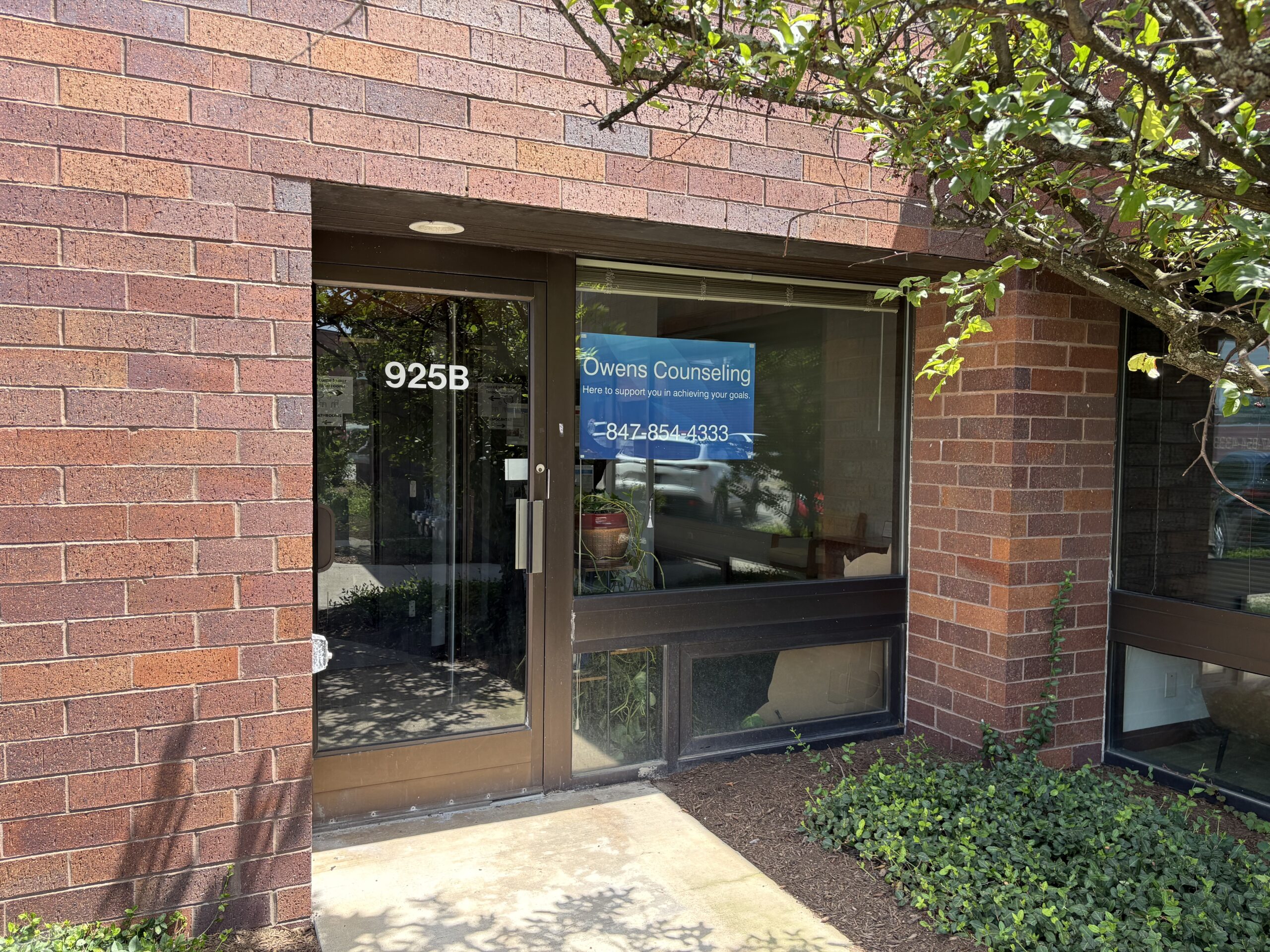 The entrance to Owens Counseling, a brick building with large windows. A teal sign above the door reads “OWENS COUNSELING.” The windows display the business name and phone number. Shrubs line the walkway outside.