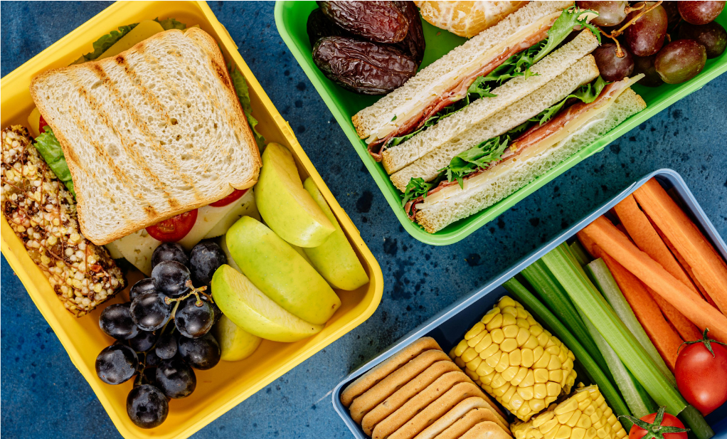 Three colorful lunchboxes contain sandwiches, sliced apples, grapes, carrot sticks, celery, corn, crackers, a granola bar, dates, and cherry tomatoes, all arranged neatly on a blue table.
