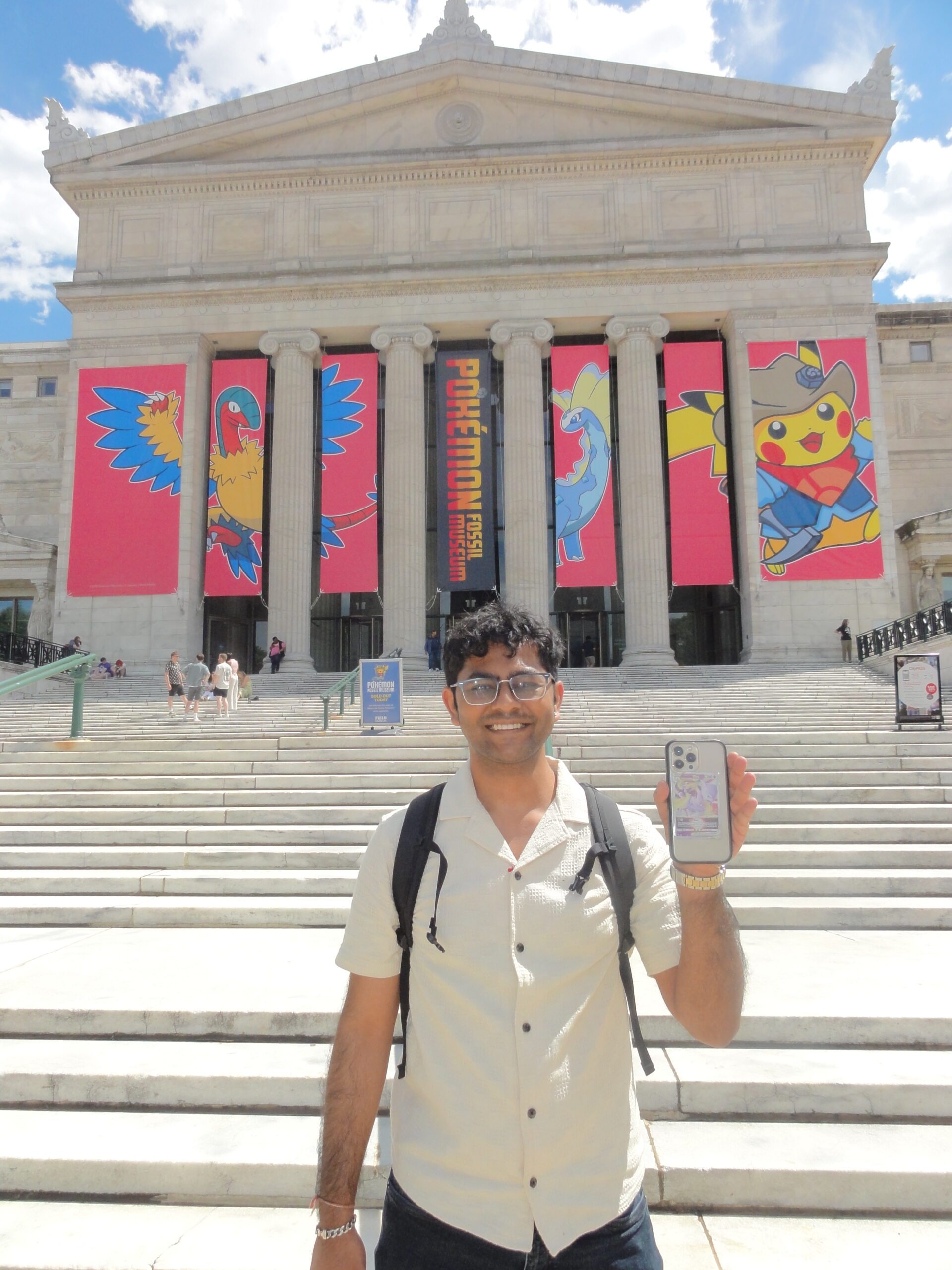 A smiling person stands on museum steps holding a phone with a Pokémon case. Large banners with Pokémon characters, including Pikachu, hang on the building behind them. It is a sunny day.
