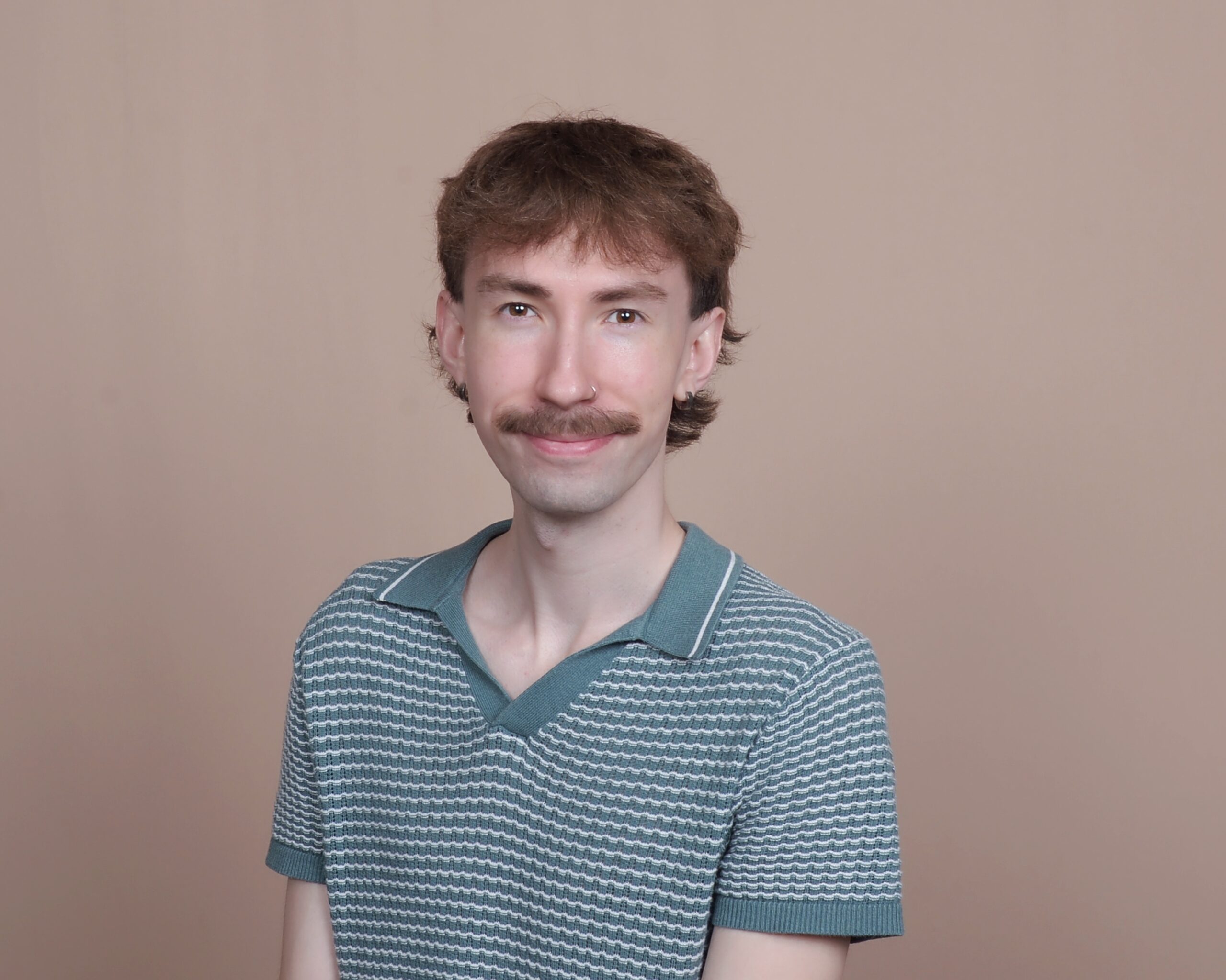 A person with brown hair and a mustache, wearing a green patterned polo shirt, smiles at the camera in front of a plain beige background.