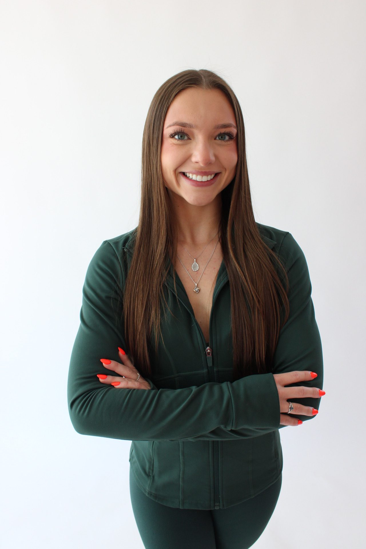 A woman with long brown hair wearing a dark green zip-up top stands with arms crossed, smiling at the camera against a plain white background. She has bright orange nails and wears layered necklaces.